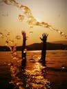 Silhouetted hands reach skyward in a golden sunset, reflecting on rippling water.
